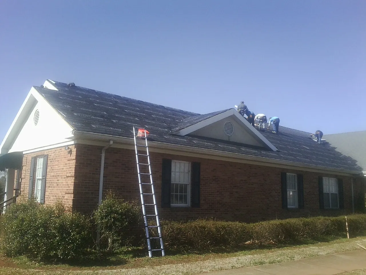 Crew tearing shingles off a brick ranch roof with a ladder set — Emergency Roof Repair work by our crew in Devol