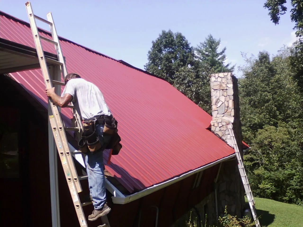 Roofer on a ladder beside a red metal roof and stone chimney — Soffit & Fascia Repair by Roofing Brothers in Devol